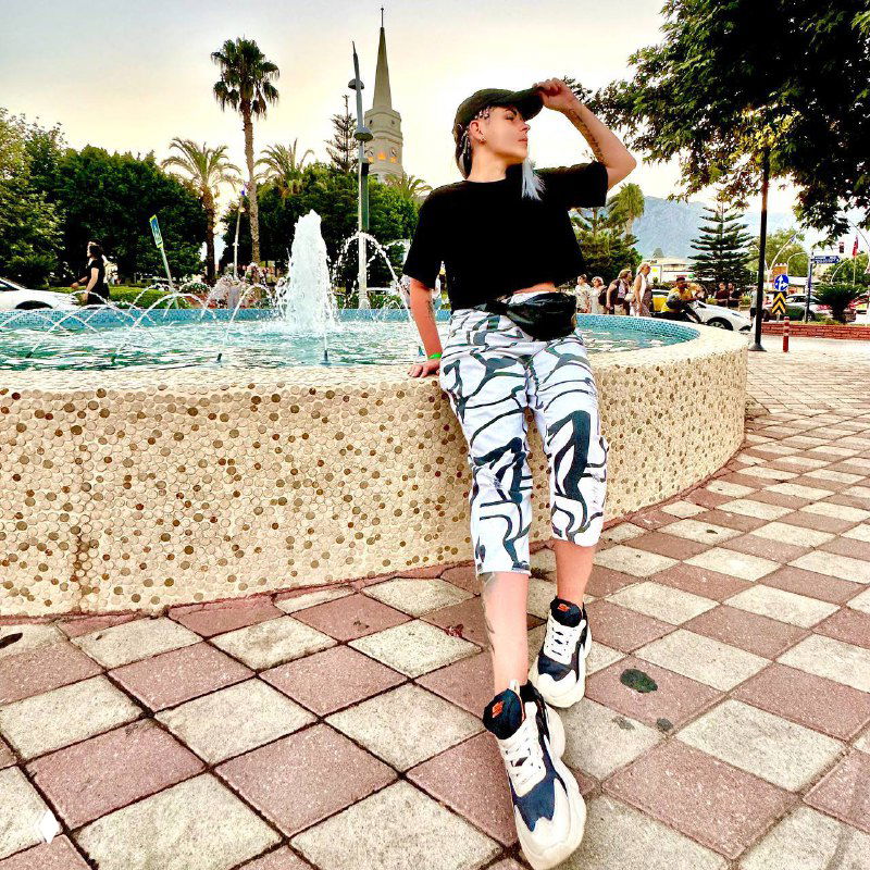 Автор канала позирует у фонтана в парке в кепке и кроссовках, повседневный образ на фоне пальм и гуляющих людей.