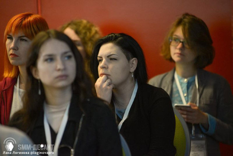 Участники конференции в зале: несколько людей с бейджами и смартфонами, сосредоточенные лица на фоне стены, деловая офлайн-атмосфера мероприятия.