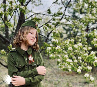 Девочка в военной форме с георгиевской ленточкой у цветущего дерева; семейное фото, подчёркивающее память, уважение и атмосферу 9 Мая
