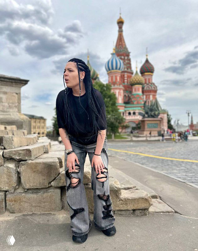 Женщина в повседневной одежде сидит на каменных ступенях на фоне собора Василия Блаженного в Москве; задумчивый образ и городской фон.