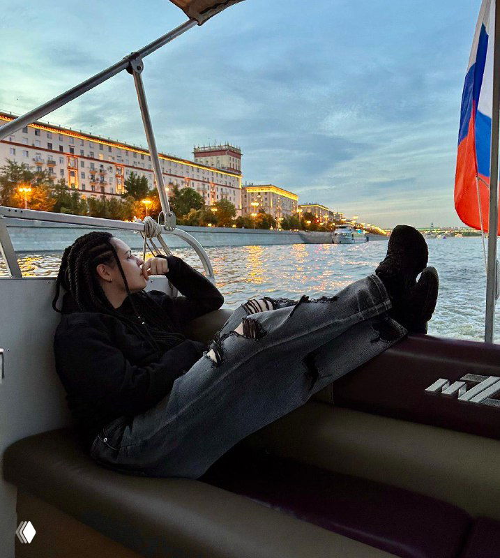 Автор сидит в кресле прогулочного катера у набережной города, отдыхает, на заднем плане видна набережная и российский флаг.