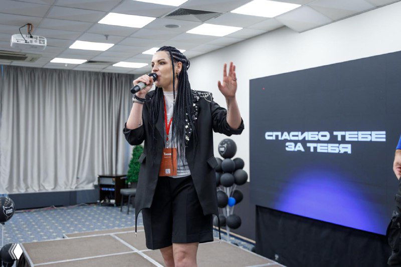 Тина выступает на сцене конференции с микрофоном; на экране за ней видно надпись «Спасибо тебе за тебя», деловой формат встречи и аудитория.