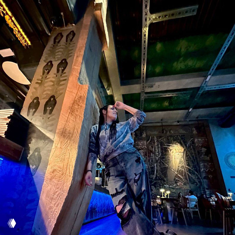 Фотография изнутри ресторана: человек опирается о декорную колонну в атмосферном зале с расписными стенами и столами, виден интерьер и посетители в фоновом плане.