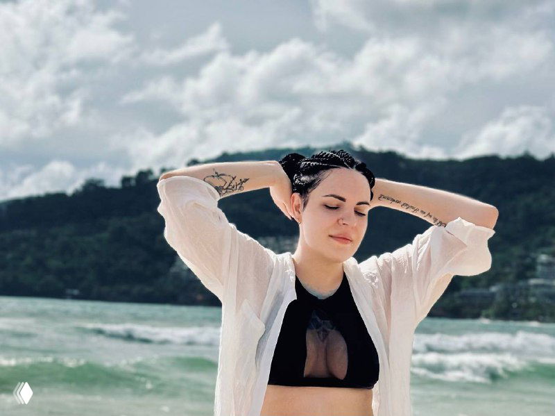 На фото Тина у моря в белой рубашке и чёрном топе, руки за головой, на заднем плане волны и низкие холмы, естественный кадр для авторского контента.