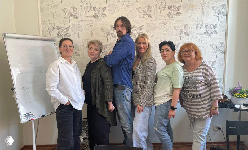 Группа сотрудников агентства стоит у флипчарта в офисе во время повышения квалификации по подбору нянь; деловая, дружелюбная атмосфера.