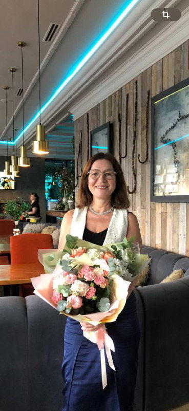 Женщина в кафе держит большой букет цветов и улыбается; портрет в тёплой интерьерной обстановке, подчёркивающий праздничное настроение и посыл поста