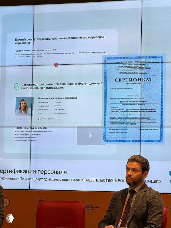 Фото экрана: слайд презентации показывает образец сертификата и данные кандидата; справа внизу сидит мужчина-докладчик.