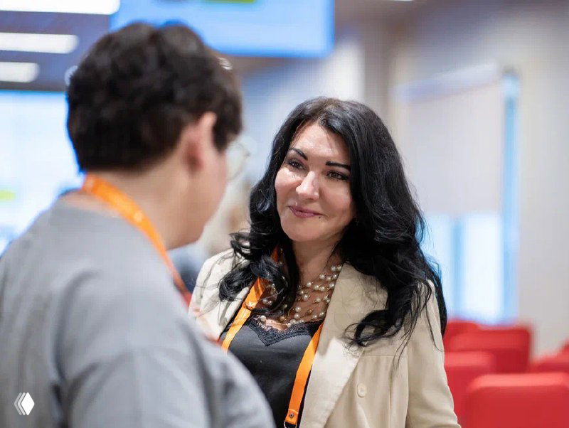 Наталья улыбается в конференционной комнате, деловой стиль одежды; кадр передаёт деловую встречу и обсуждение проектов по подбору нянь и сертификации.