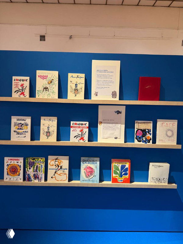 Фрагмент экспозиции: несколько детских книжных обложек и миниатюр на полке на фоне синей стены в музее, вид крупным планом.