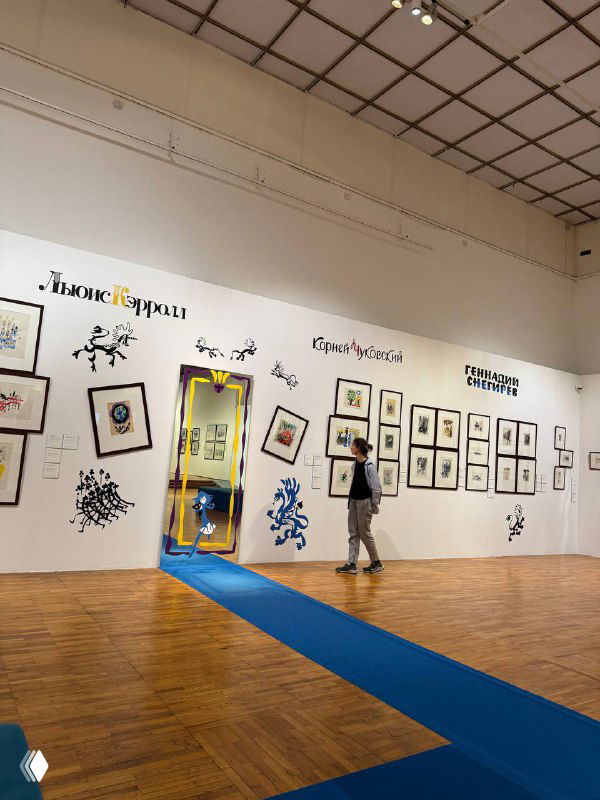 Широкий кадр зала Новой Третьяковки с оформленной стеной иллюстраций, посетитель проходит мимо экспозиции на синем напольном ковре.