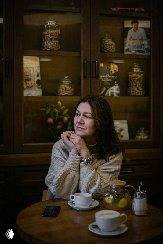 Светлана Тошич в уютном кафе за столом с чашками, задумчиво улыбается; на заднем плане полка с банками и книгами, создавая контекст беседы о подборе нянь.