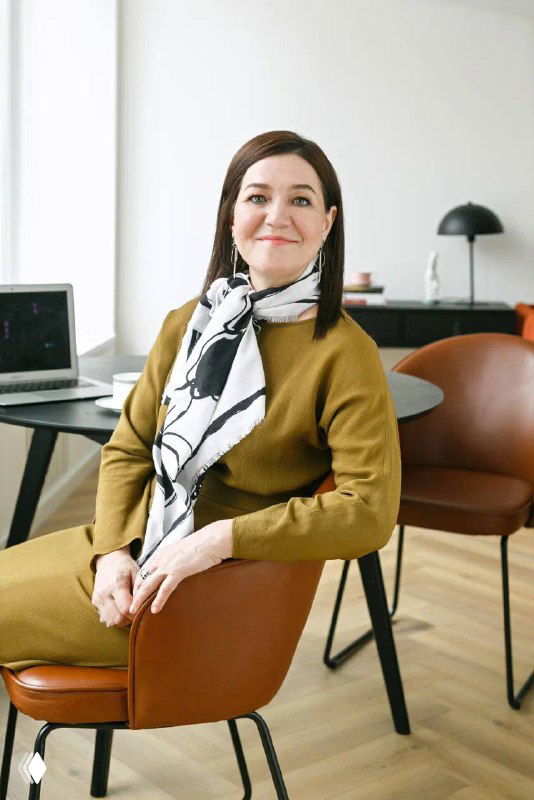 Портрет улыбающейся женщины в офисе, сидит в кресле на фоне стола и ноутбука — иллюстрация к статье о рынке нянь.