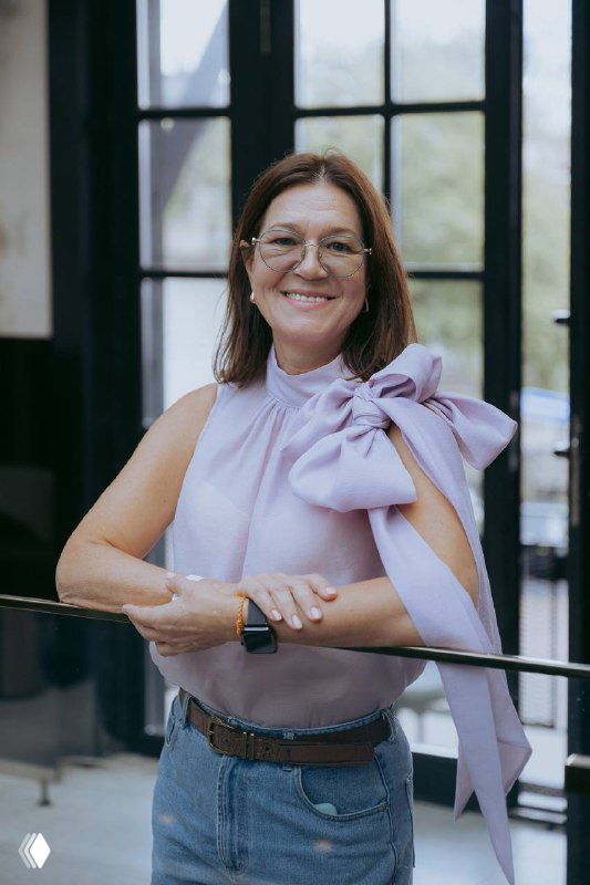 Портрет улыбающейся женщины в светлой блузе с большим декоративным бантом, стоящей у окна в интерьере; нейтральный фон, доброжелательный образ для темы ухода за ребёнком.