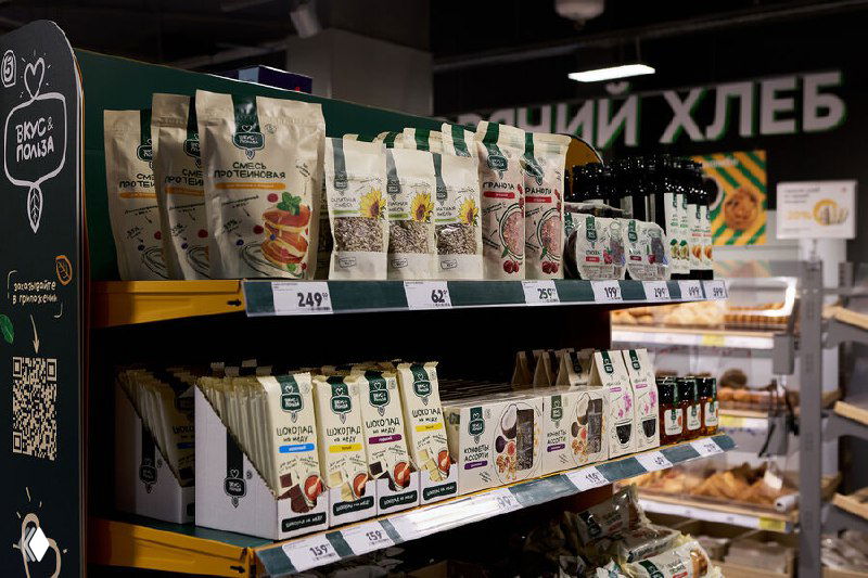 Полка магазина с упакованными цельнозерновыми продуктами и хлебцами на фоне отделения выпечки — пример расширяющегося ассортимента ЗОЖ-продуктов на полках ритейла.