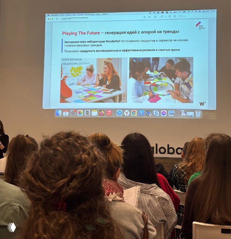 Зал с участниками тренинга и проекторной презентацией метода Playing the future: люди смотрят на слайды и обсуждают задания в группах.