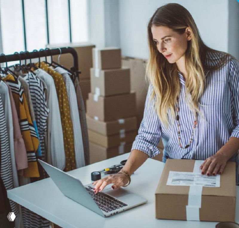 Женщина у ноутбука в интернет-магазине упаковывает заказ в коробку на фоне одежды — иллюстрация онлайн‑розницы и логистики.