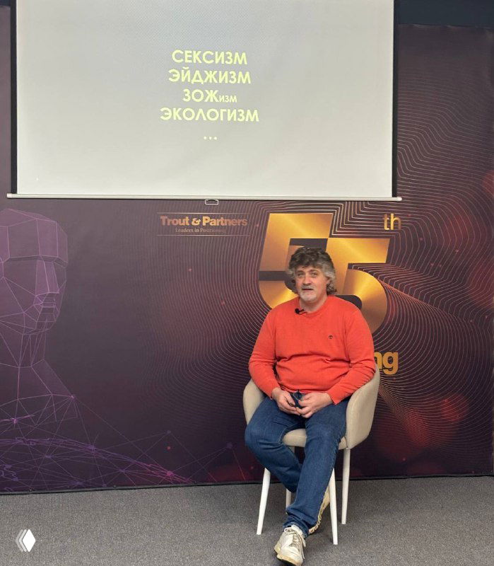Святослав Петров сидит на стуле на сцене конференции, на проекции надписи «СЕКСИЗМ ЭЙДЖИЗМ ЗОЖизм ЭКОЛОГИЗМ» и элемент брендинга мероприятия.