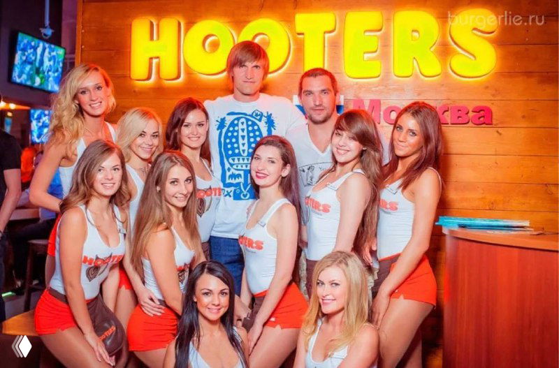 Группа официанток сети Hooters в фирменной форме у неонового логотипа Hooters в интерьере ресторана — иллюстрация концепции бренда.