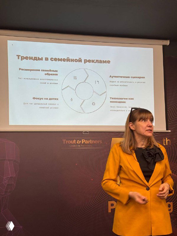 Женщина в жёлтом пиджаке выступает с докладом; на экране позади диаграмма трендов семейной рекламы и ключевые тезисы.