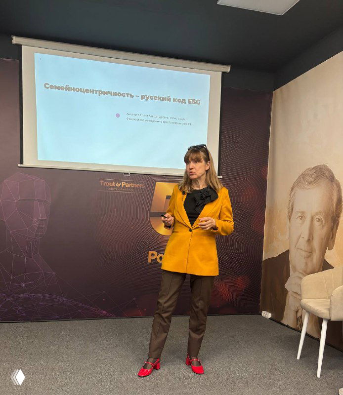 Спикер у экрана с заголовком о семейноцентричности; сцена мероприятия и крупный портрет в оформлении зала рядом.