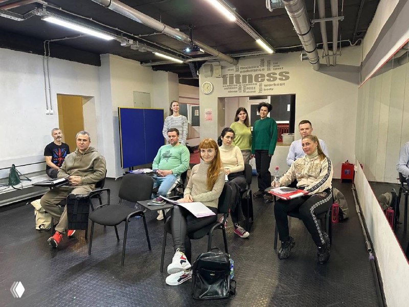 Очное занятие в Академии Светланы Алаевой (Тверь): группа будущих фитнес‑тренеров в зале, сидят на стульях с тетрадями и сумками, готовые к учебе.