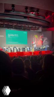 Фото конференции: затемнённый зал, сцена с презентацией на зелёном слайде и красной подсветкой, силуэты аудитории на переднем плане.