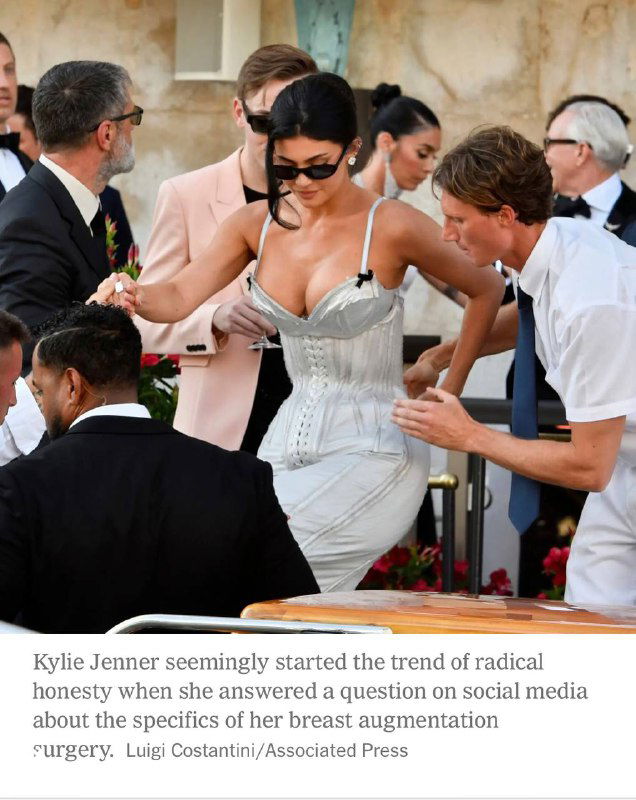 Женщина в белом корсете на светском мероприятии: ассистенты помогают поправить платье; кадр подчёркивает тему пластики и публичности.