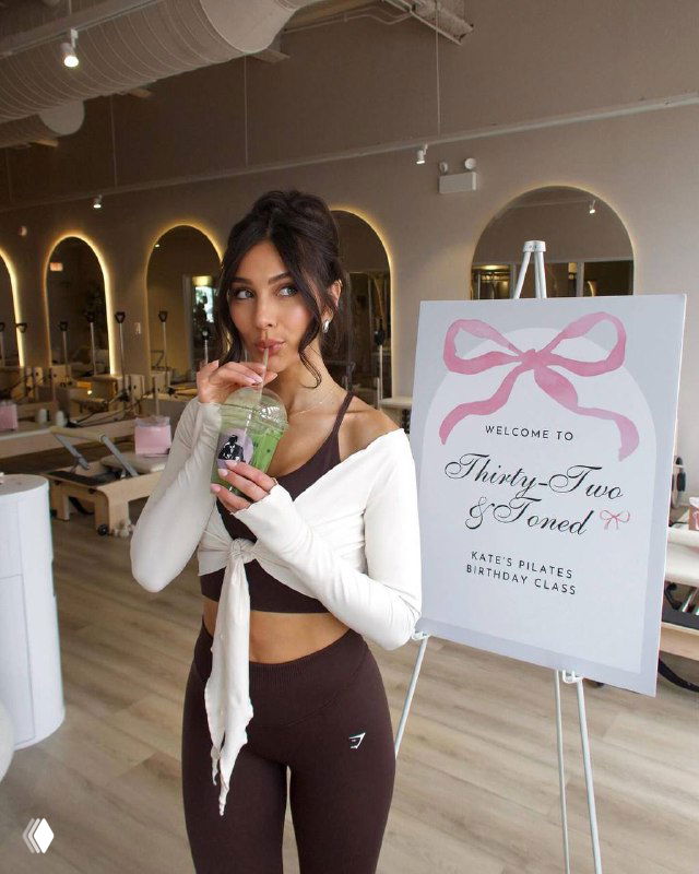 Женщина в светлом спа‑пространстве с напитком рядом с рекламным стендом мероприятия — пример спроса на элитные wellness‑ивенты.