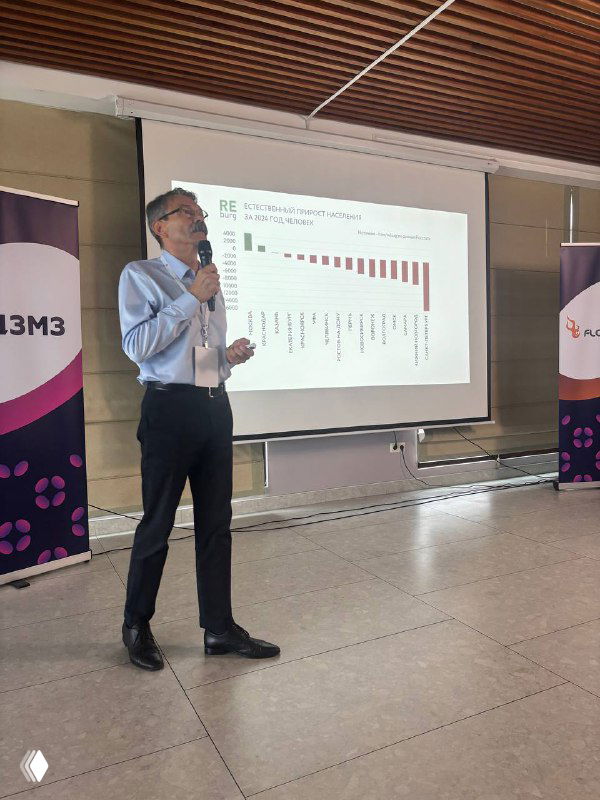 Докладчик на сцене конференции по митохондриальной и превентивной медицине; человек в деловой одежде стоит перед экраном с графиком и логотипами организаторов.