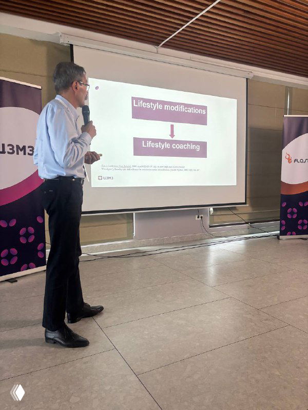 Докладчик у экрана с слайдом, где видны слова «Lifestyle medicine / Lifestyle coaching», конференц-зал и брендовые баннеры по бокам.