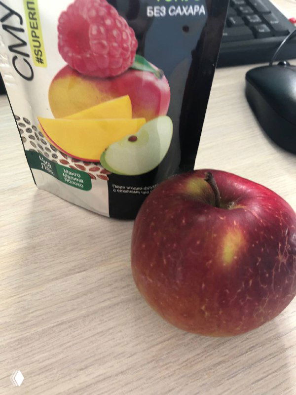 Красное яблоко на рабочем столе рядом с упаковкой фруктового напитка с пометкой «без сахара»; на заднем плане видна клавиатура и компьютерная мышь.