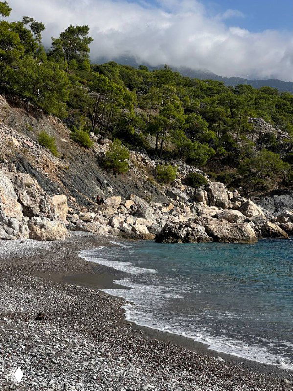 Побережье с кромкой прибоя на гальке, спокойная морская гладь и лесистые склоны вдалеке — детали пляжного берега Ликийки весной или осенью.