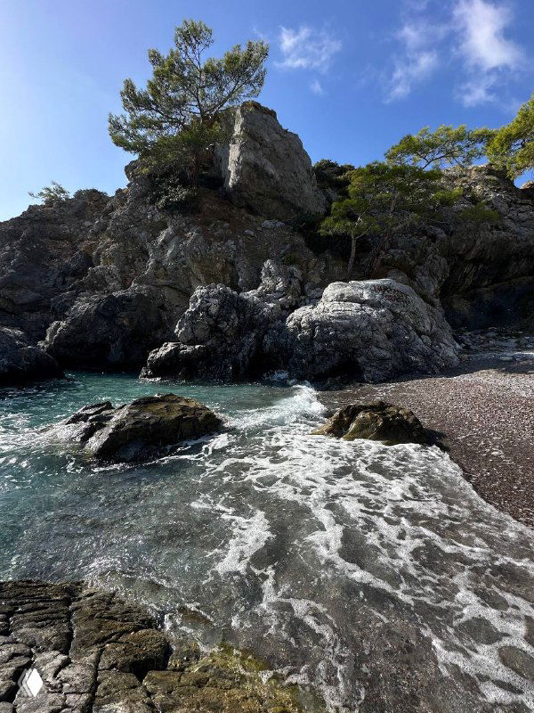 Скалы у воды с волнами и морской пеной, темные камни и прибрежная растительность — драматичный фрагмент побережья рядом с пляжем.