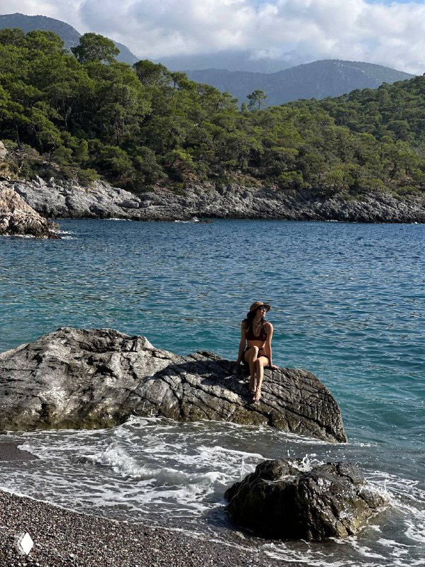 Человек сидит на крупном камне у моря, вид на спокойную бухту и горы на фоне — момент отдыха после похода на берегу Ликийки.