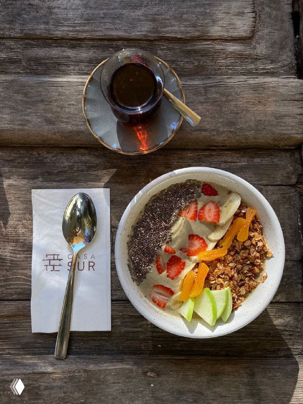 Смузи-боул с фруктами и гранолой на деревянном столе рядом с чашкой чая и салфеткой с логотипом Casa Sur — завтрак в саду.