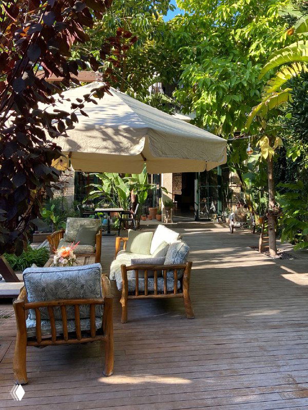 Уютная зона с низкой мебелью и зонтами на деревянной террасе Casa Sur: мягкие кресла, цветущие растения и много тени под деревьями.