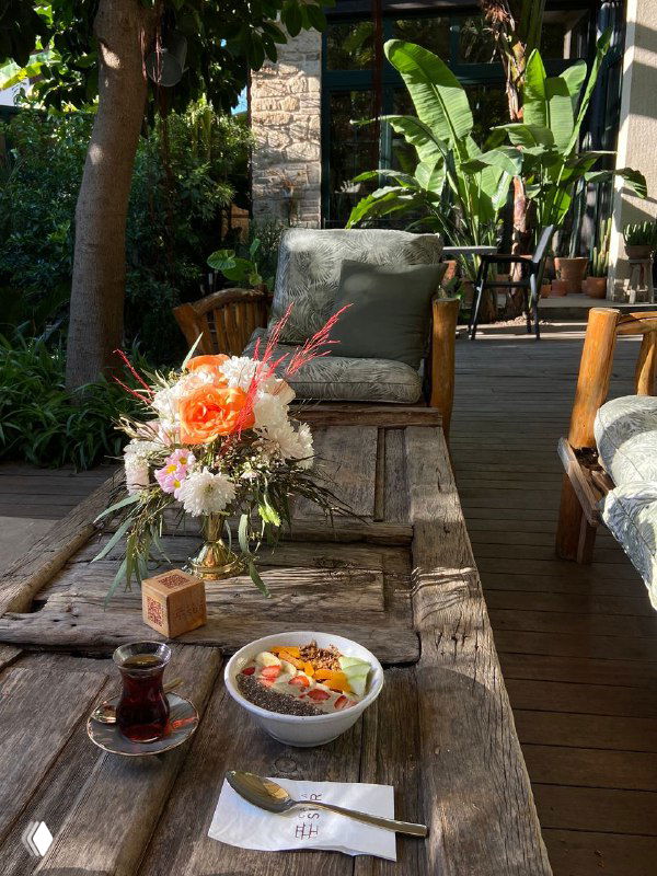 Деревянный стол с букетом цветов, чашкой чая и смузи-боулом на фоне зелёной обивки и садовой мебели в Casa Sur — атмосферный завтрак.