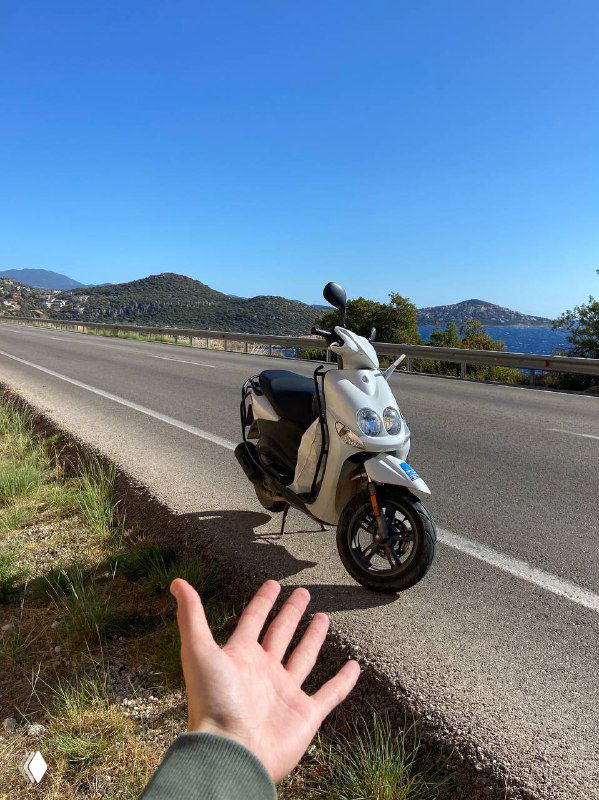 Скутер припаркован на обочине прибрежной трассы, на переднем плане рука автора, на заднем плане дорога и море — момент перед стартом в путешествии.