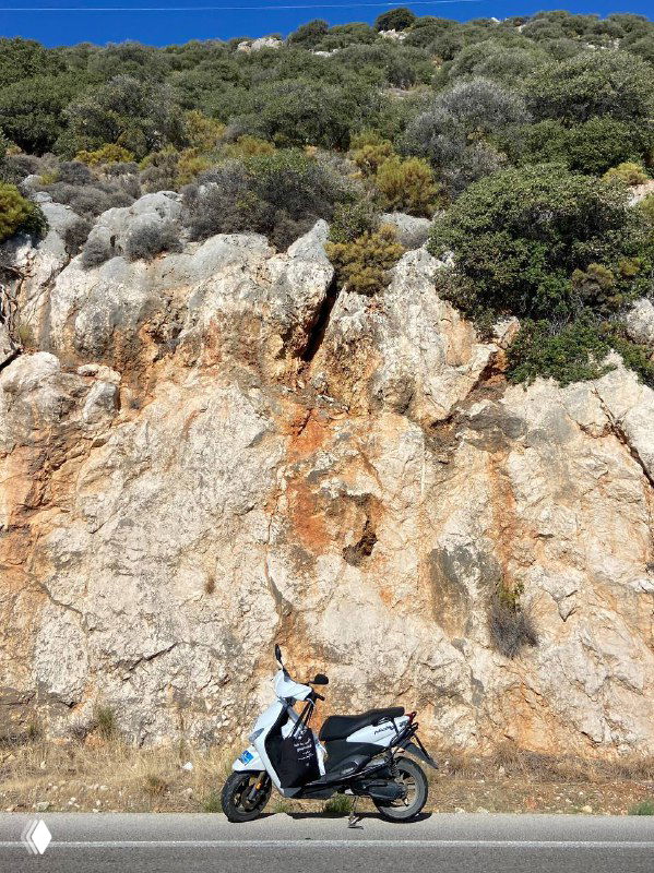 Скутер припаркован у основания каменистого обрыва, на фоне видна сухая растительность и скалистый склон — фото на маршруте вдоль побережья.