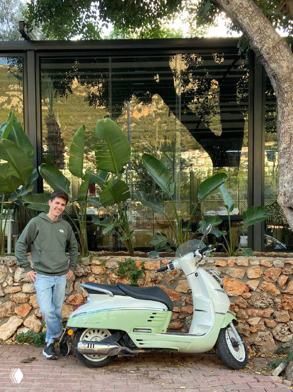 Светло-зелёный ретро-скутер у стеклянного фасада кафе, рядом автор в худи — финальный кадр аренды и атмосферного дня на островке побережья.