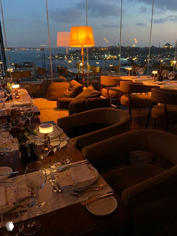 Интерьер ресторана на крыше вечером с сервированными столами у панорамных окон и видом на Босфор, огни города и корабли.