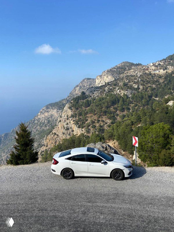 Белый автомобиль припаркован на узкой горной дороге без ограждений, вокруг скалы и обрыв, видна часть побережья и ясное небо.