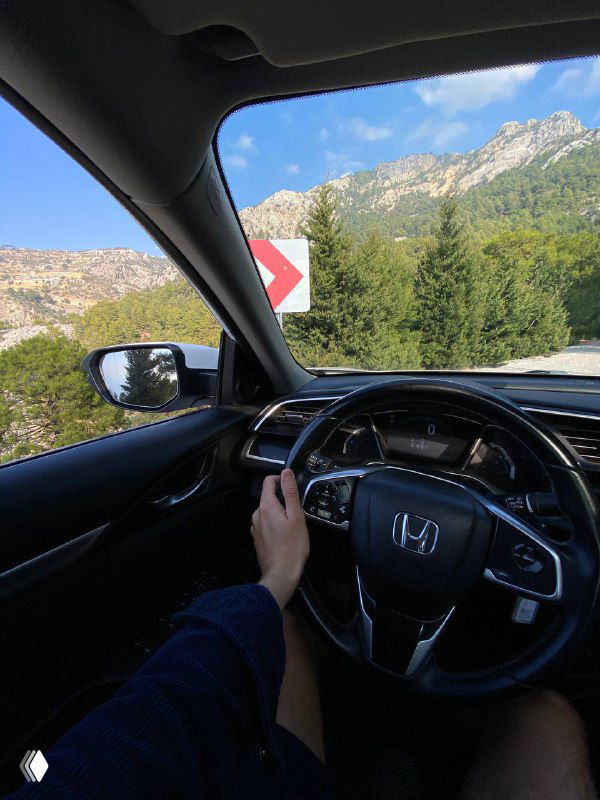 Руки водителя на руле внутри автомобиля, через лобовое стекло видна извилистая горная дорога, скалы и зеленые склоны — поездка по серпантину.