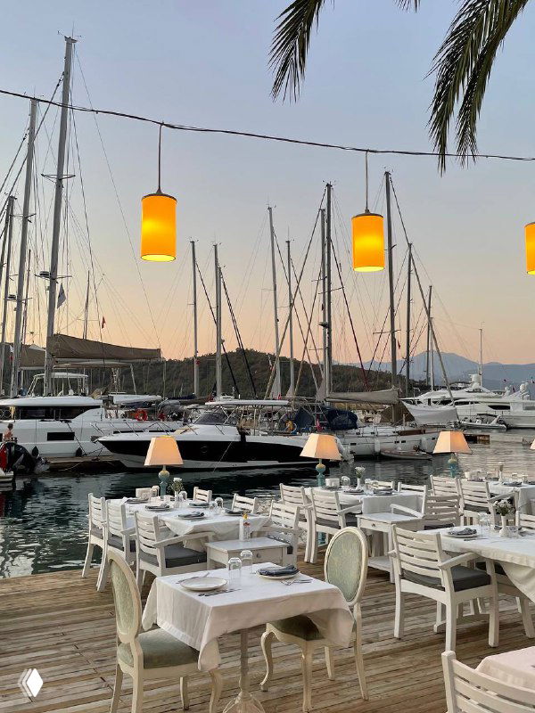 Терраса Mori Restoran у марина: белые сервированные столы и стулья, лампы над столиками, пришвартованные яхты и горы на закате.