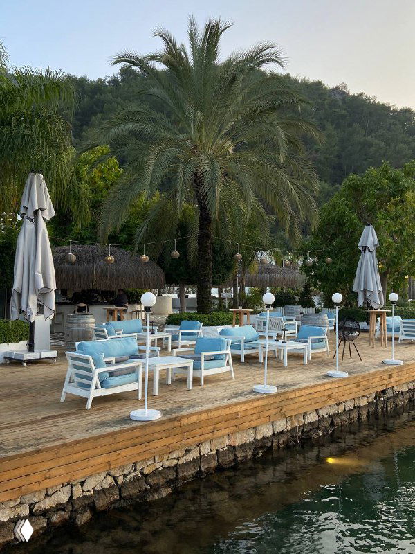 Набережная и зона отдыха Mori Restoran: голубые диваны и белая мебель у воды, пальмы и зелёные холмы на фоне курорта Фетхие.