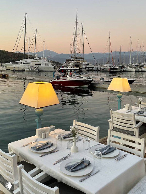 Ужин у причала в Mori Restoran: сервировка стола на фоне марина, настольные лампы, яхты и спокойная водная гладь в лучах заката.