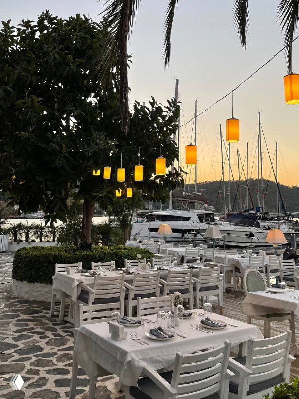 Вечерняя терраса Mori Restoran: ряд белых столов вдоль мачт яхт, тёплое освещение подвесных ламп и пейзаж марина на закате.