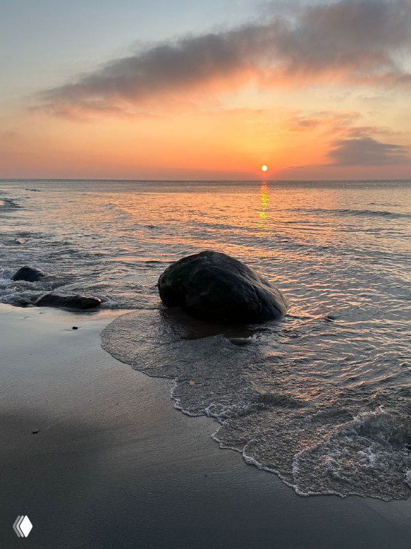 Закат над Балтийским морем у берега Калининградской области: спокойная вода, крупный камень на песчаном пляже и тёплая пастельная палитра неба.