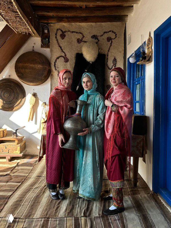 Три женщины в традиционных дагестанских нарядах внутри этно‑сакли среди керамики и текстиля, музейная атмосфера дома хозяев.
