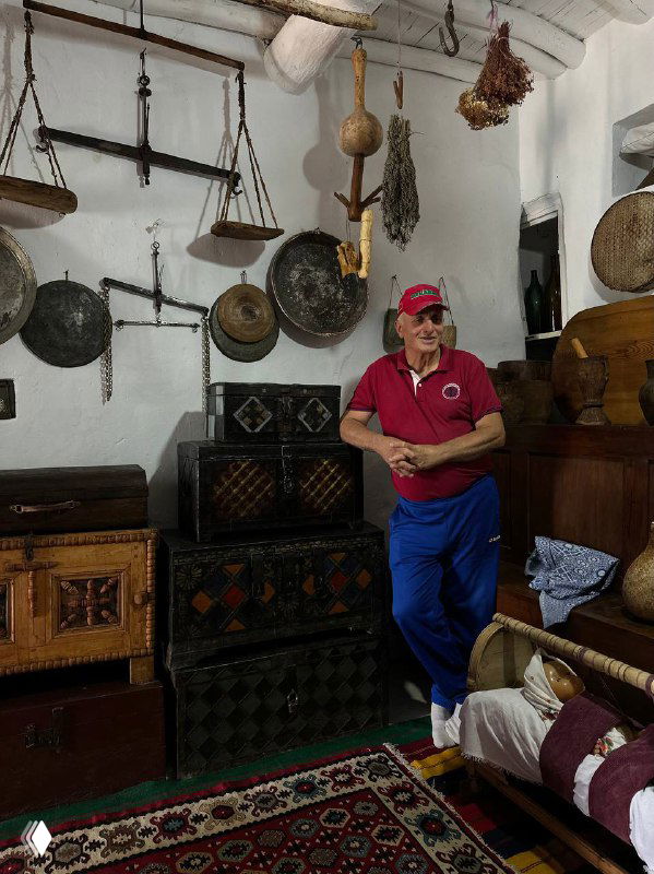 Магомед в комнате с коллекцией старинных предметов: музыкальные инструменты, сундуки и деревянный интерьер этно‑сакли.